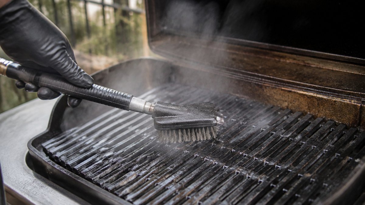 Steam cleaning brush scrubbing grease off warm metal grill grates with visible steam rising
