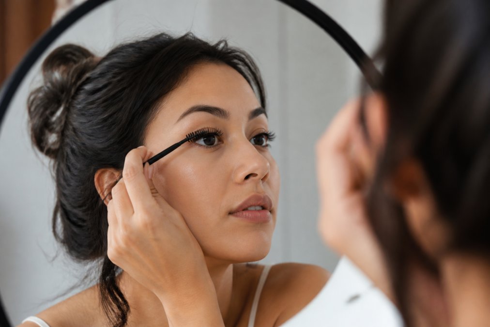 Close-up of a woman gently brushing her eyelash extensions with a clean spoolie wand in front of a bathroom mirror, soft natural lighting, her face relaxed and focused