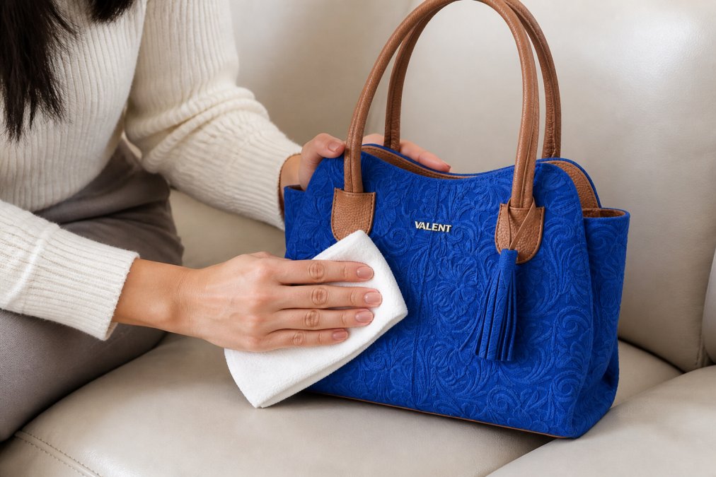 Close-up of hands gently wiping a leather handbag with a soft microfiber cloth on a light-colored leather surface, showing proper cleaning technique in natural daylight