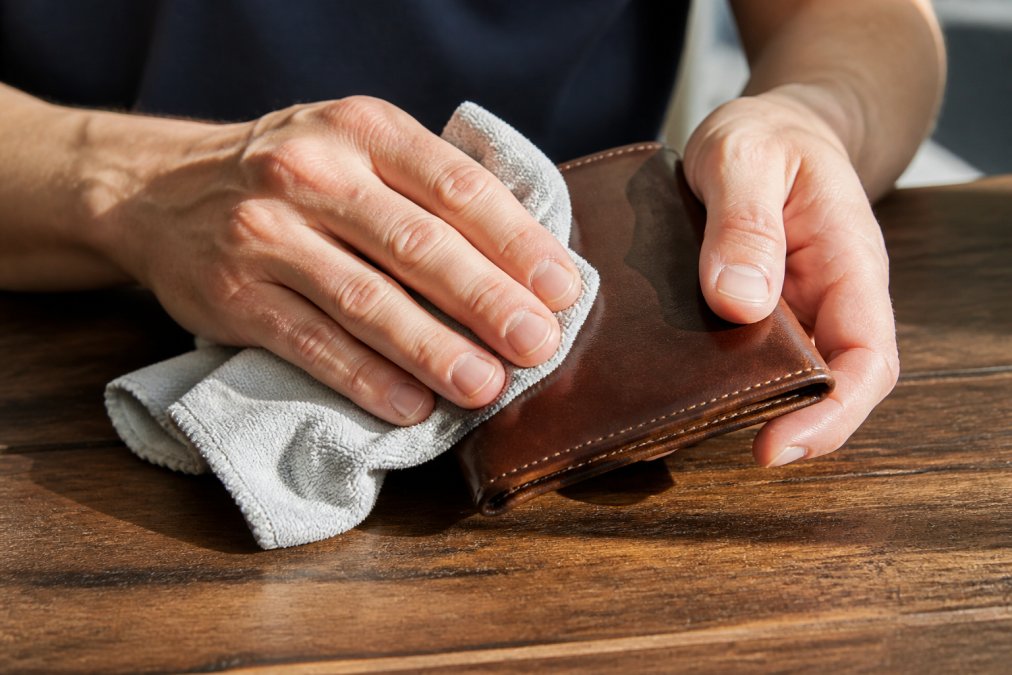 Close-up of a person's hands gently wiping a brown leather wallet with a soft microfiber cloth on a clean wooden table, natural window light casting soft shadows