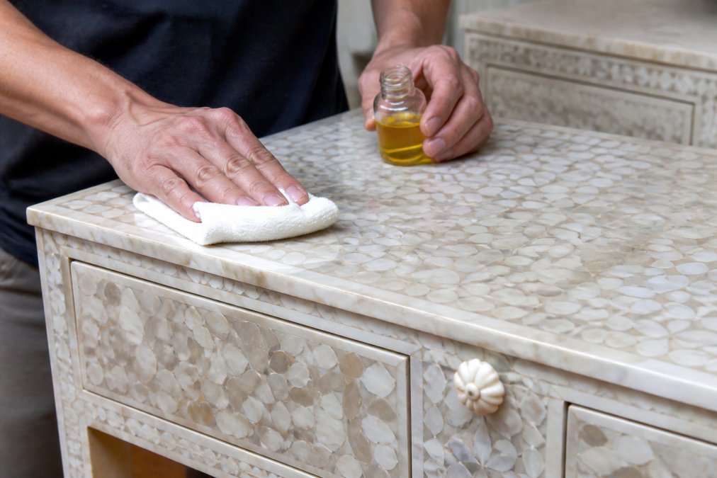 Hands applying mineral oil to a handcrafted mother of pearl inlay furniture piece using a soft cloth, demonstrating gentle polishing and restoration technique with natural diffused lighting