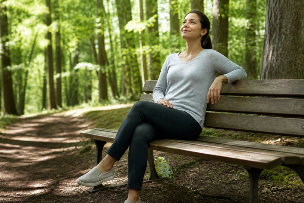 Adult sitting peacefully on a wooden bench in a natural outdoor setting, trees and soft natural light in background, with a calm and content expression, representing mental clarity and stress reduction