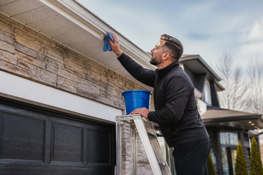 Homeowner cleaning a permanent holiday lights track under a house soffit with a microfiber cloth and soapy water