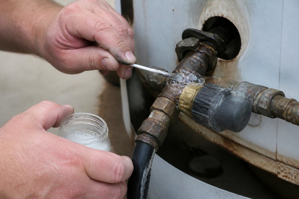 Close-up of hands applying soapy water solution to propane appliance connection points with a small brush, showing bubbles forming around a gas leak on a metal fitting in natural daylight