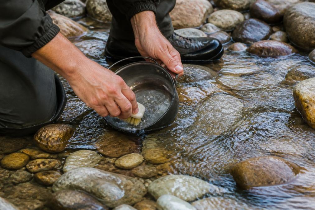 Hands washing a camping cookware pot in a stream with a soft brush, showing proper field cleaning technique with natural daylight on water