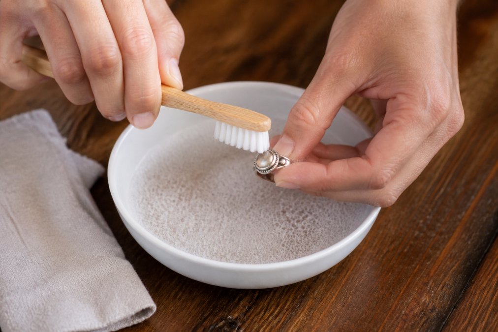 Close-up of hands gently scrubbing a 925 sterling silver ring with a soft-bristled toothbrush and soapy water in a small white bowl on a wooden table, with a lint-free cloth nearby