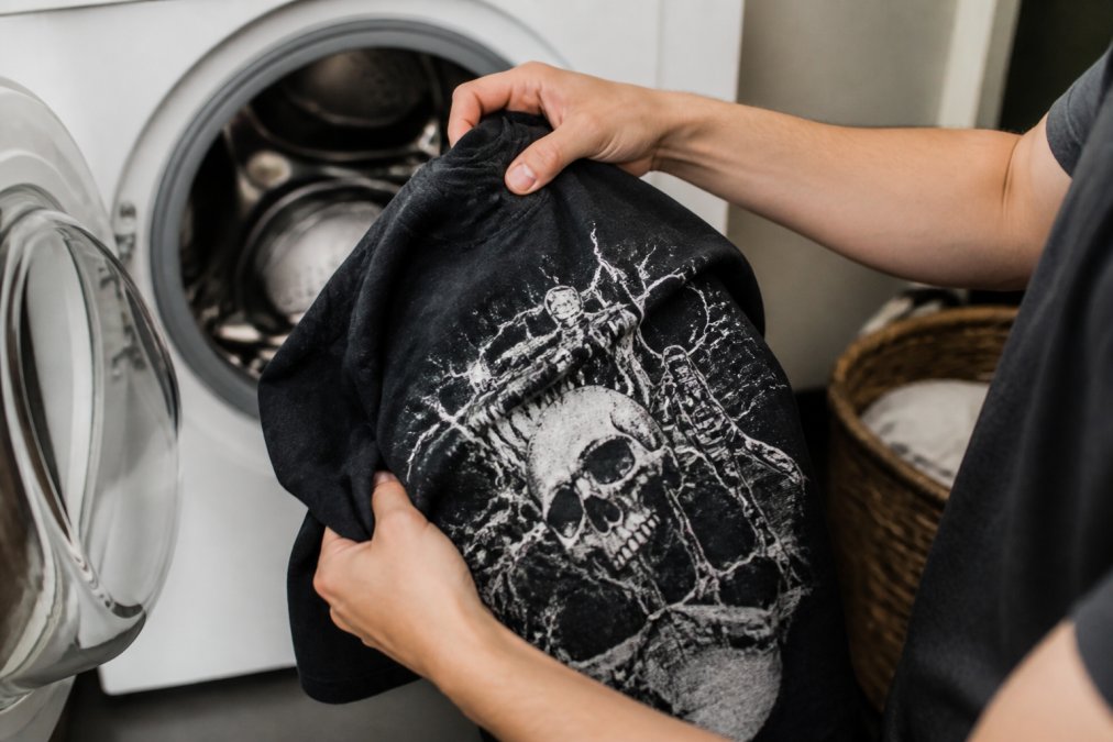 Close-up of hands turning a graphic tee inside out before placing it in a washing machine, showing the detailed print on the back