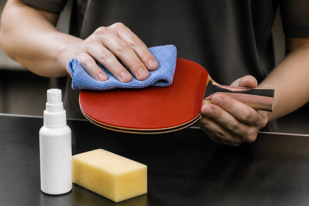 Close-up of hands using a microfiber cloth to gently wipe the rubber surface of a table tennis racket, with a bottle of rubber cleaner and a sponge nearby on a clean table