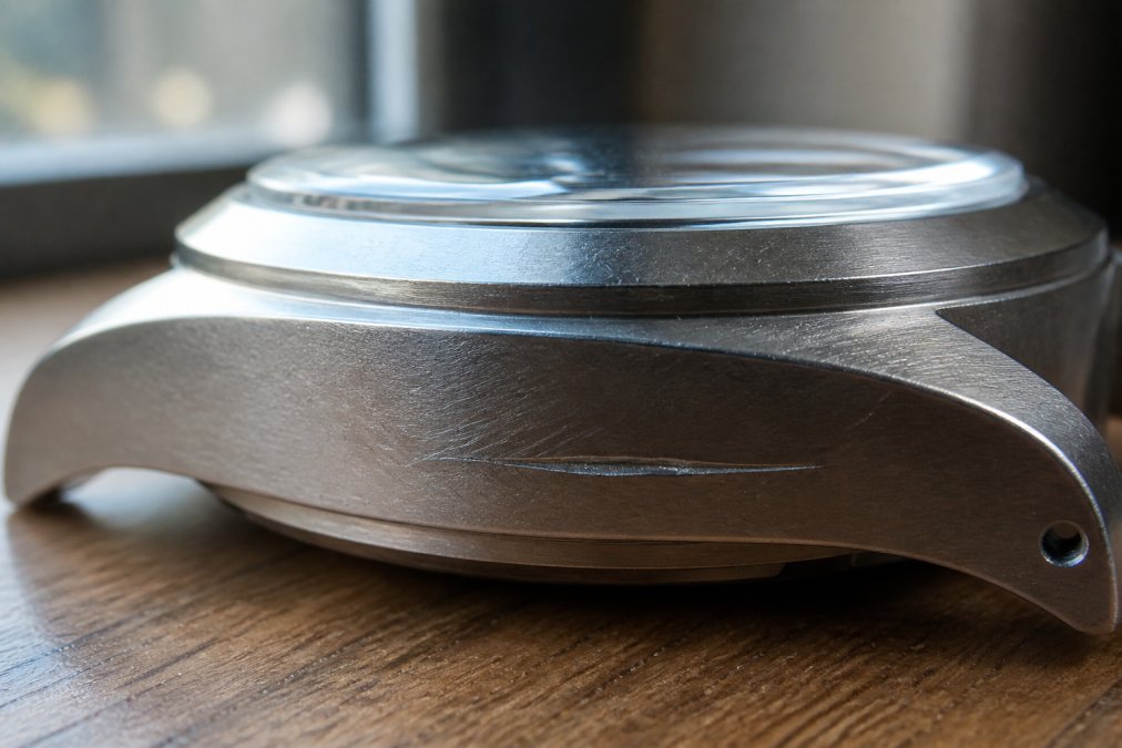 Close-up of a titanium watch case surface showing the contrast between light surface scuffs that catch the light and a deeper scratch that creates a visible groove in the metal, photographed under natural window lighting