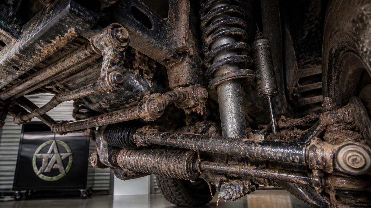 Close-up of a truck's undercarriage and suspension components showing shocks, bushings, and steering linkage with visible mud and trail debris
