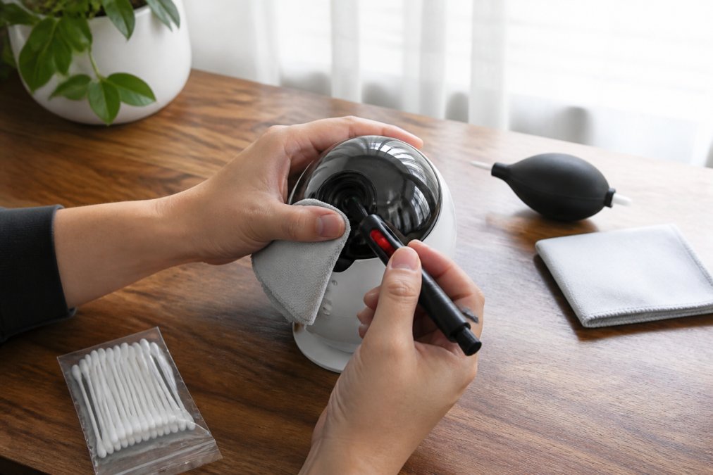 Close-up of a person's hands using a microfiber cloth and a cleaning pen to gently wipe the lens of a small dome-shaped galaxy projector on a wooden desk, soft daylight from a window