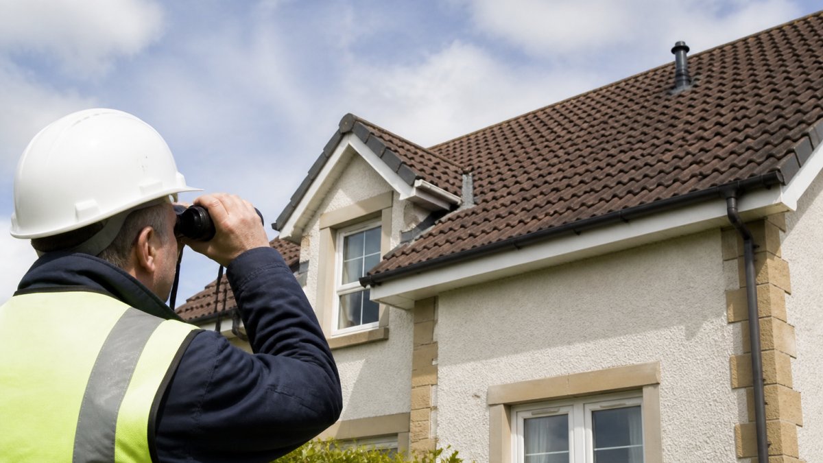 Professional roofer safely inspecting residential roof from ground level using binoculars, with clear visibility of roof tiles, flashing, and guttering in natural daylight