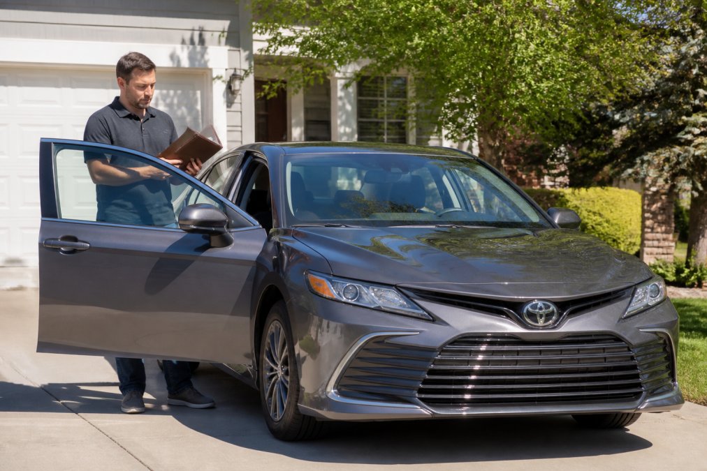 A clean, late-model Toyota sedan parked in a bright suburban driveway at midday, the owner standing beside the open driver's door reviewing a leather service folder