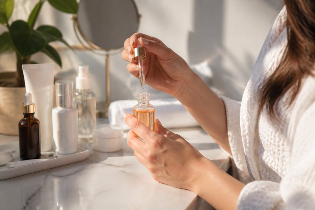 Close-up of a woman's hands holding a small glass serum bottle with a dropper, with skincare products arranged neatly on a white marble surface in soft morning light