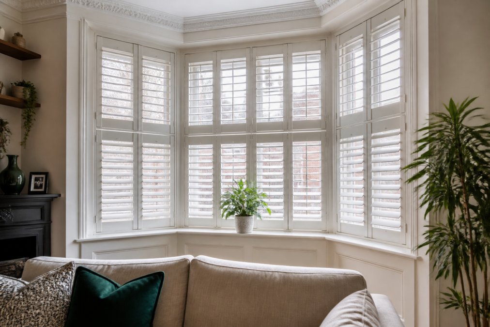 A bright Victorian bay window in a London living room fitted with white made-to-measure plantation shutters, slats partially tilted to show light control and privacy, a plant on the sill and a sofa visible in the foreground
