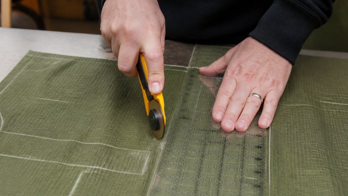 Close-up of hands using rotary cutter and ruler on ripstop nylon fabric with chalk markings visible on work table