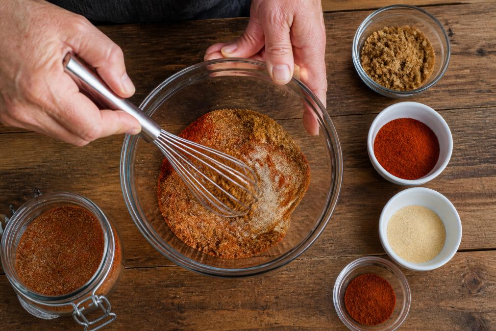 Hands whisking brown sugar and spices in a glass bowl to prepare a homemade sweet and spicy rub on a wooden counter.