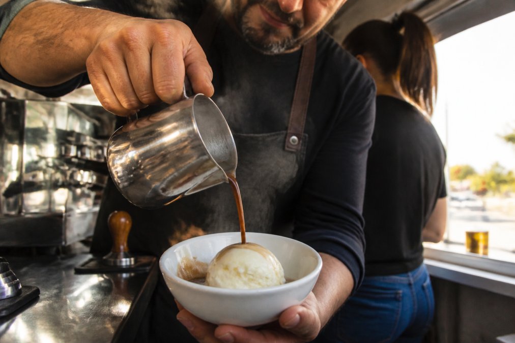 Close-up of hot espresso being poured over a scoop of vanilla bean gelato in a white ceramic bowl, steam rising visibly, with the gelato beginning to melt slightly at the edges where the espresso makes contact