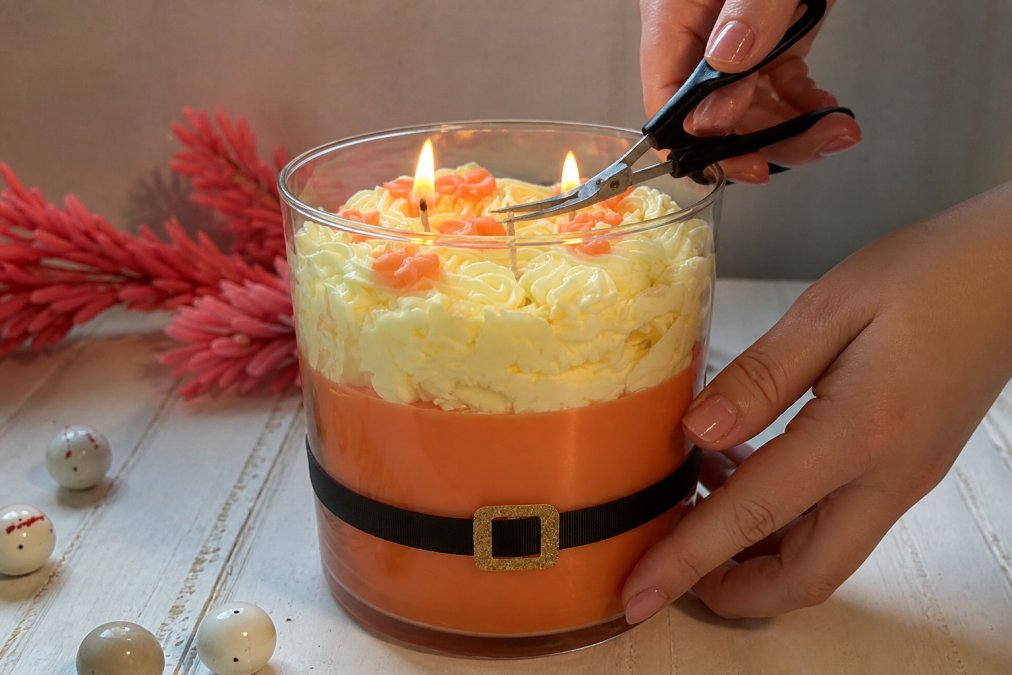Close-up of hands using small scissors to trim a candle wick to 1/4 inch length above the wax surface, with a lit candle visible in soft natural lighting
