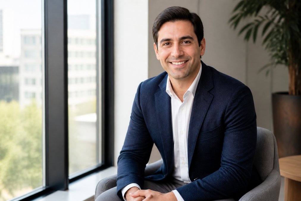A business professional in a navy blazer seated near a large floor-to-ceiling window, soft diffused natural daylight illuminating one side of their face with a gentle fill on the other, creating an approachable and relaxed corporate portrait in a modern office setting