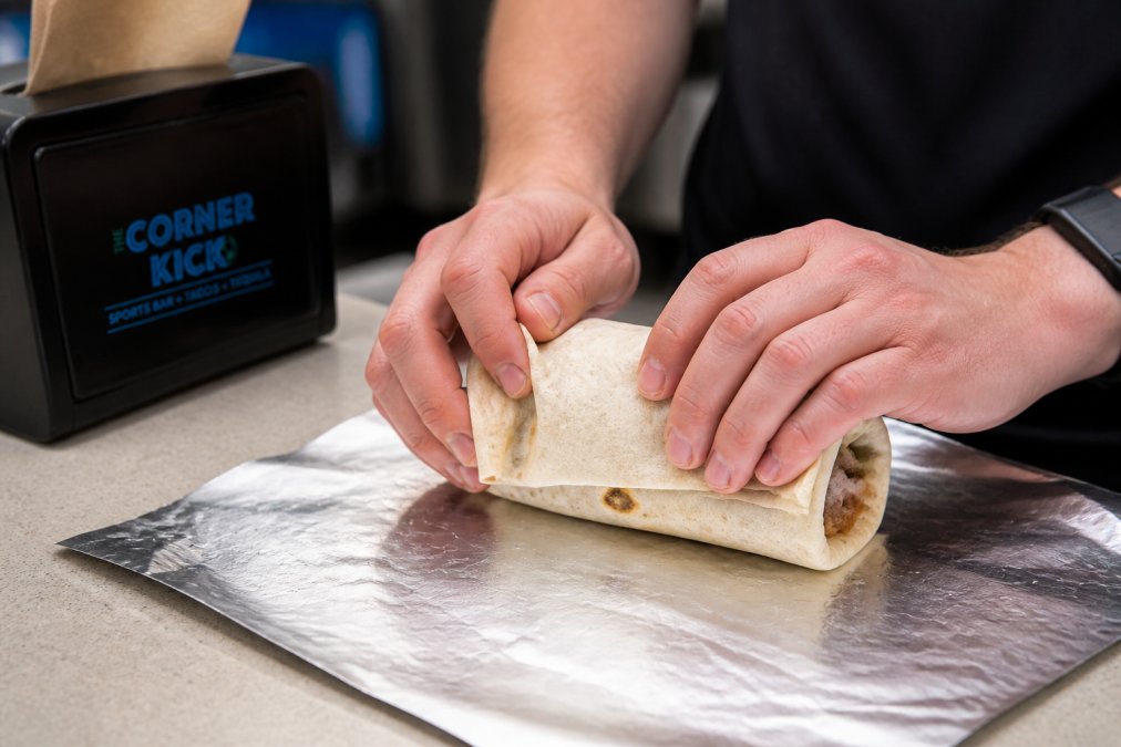 Close-up of hands demonstrating the tight roll technique, showing the burrito being wrapped with the edges tucked in, with a foil wrapper partially visible