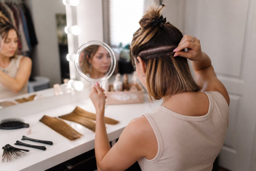 Woman with a short bob clipping wefts into her hair to make extensions look natural at a vanity mirror.