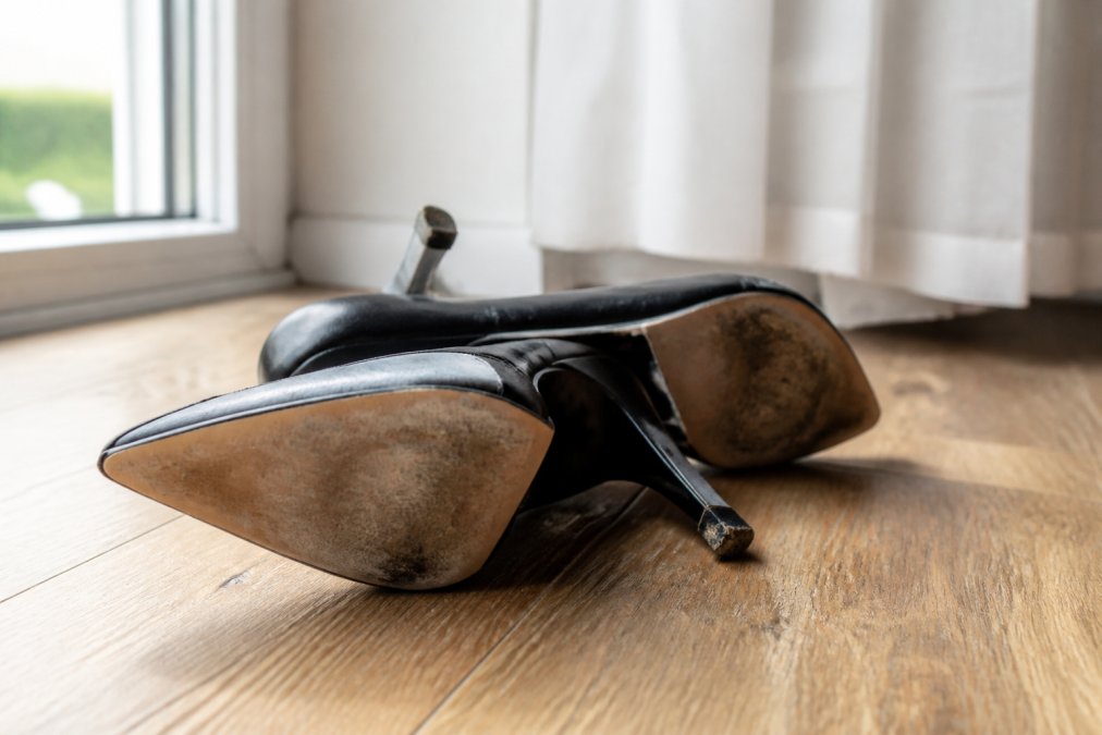 A pair of elegant black high heels on a wooden floor showing visible wear on the heel tip and scuffed sole, soft natural light streaming from a nearby window