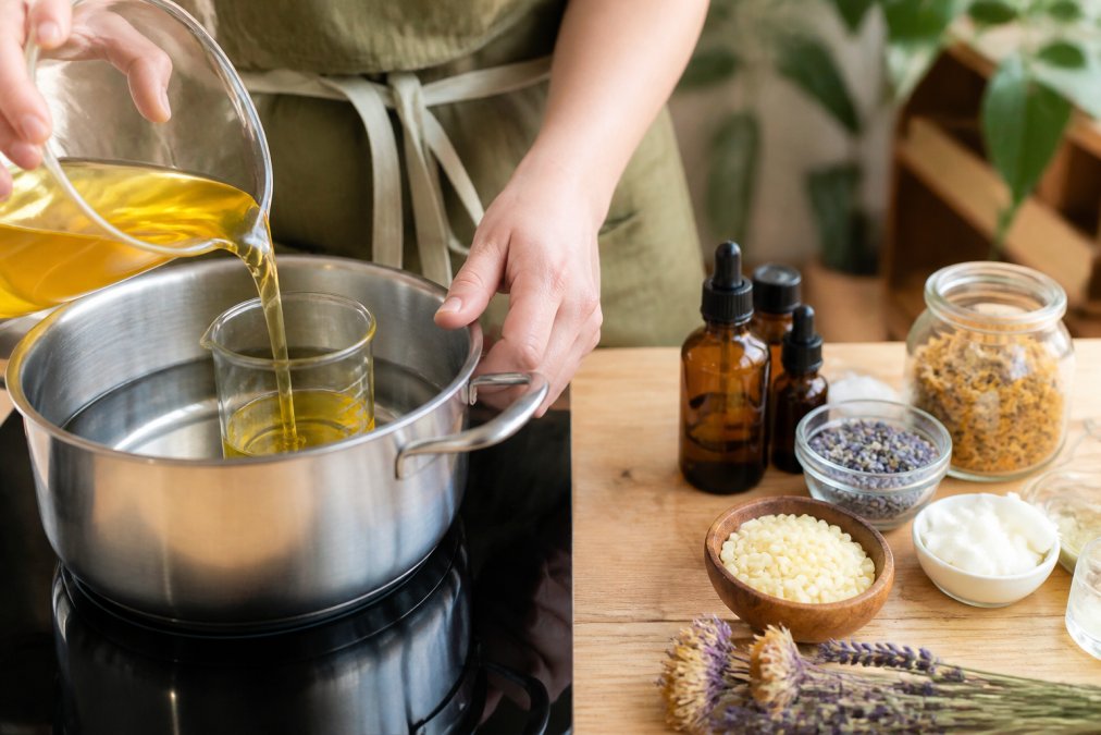 Close-up of hands pouring melted beeswax and carrier oil into a glass container over a double boiler setup with essential oils and botanical ingredients visible nearby