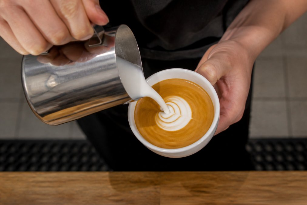 Close-up overhead shot of hands tilting a stainless steel milk pitcher while pouring steamed milk into a white ceramic cup of espresso, with a heart pattern beginning to form on the surface