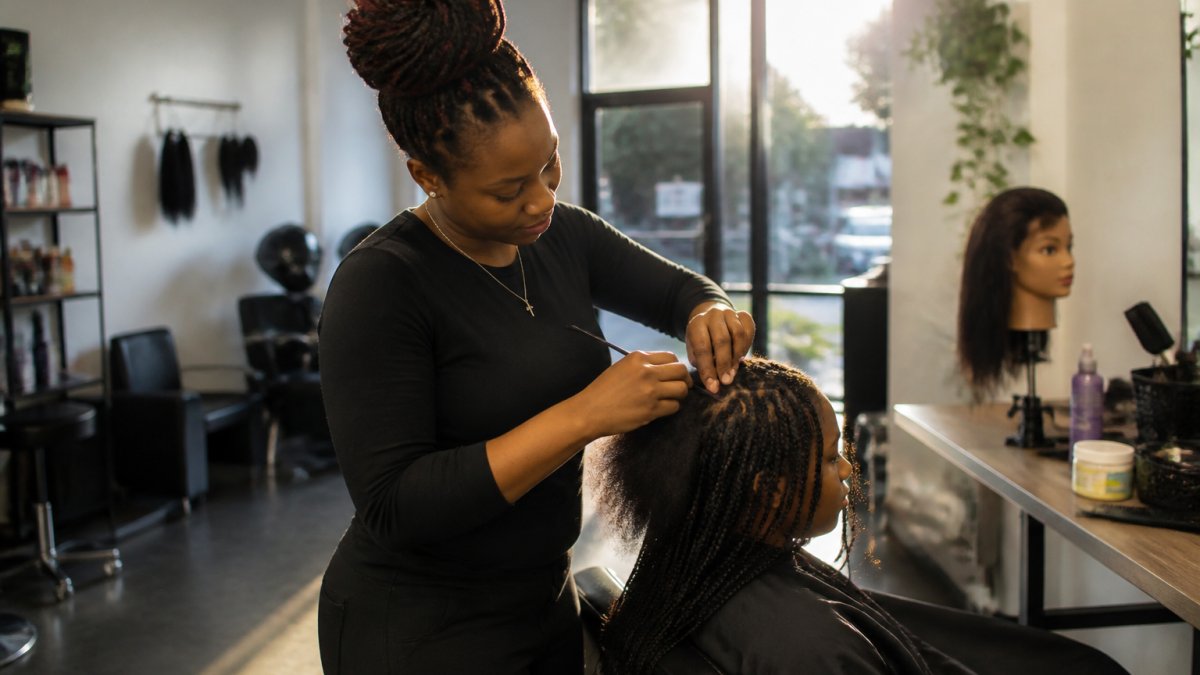 A close-up of a braider's hands carefully parting and braiding a client's hair in a bright, welcoming salon studio, with a mannequin head and practice tools visible in the background