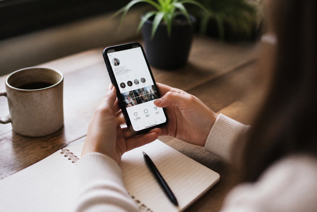Person holding smartphone showing Instagram app with engagement metrics visible on screen, sitting at desk with coffee and notebook