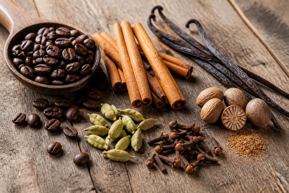 Close-up of whole spices including cinnamon sticks, cardamom pods, cloves, and nutmeg arranged next to whole coffee beans and a vanilla bean on a weathered wooden surface with natural morning light