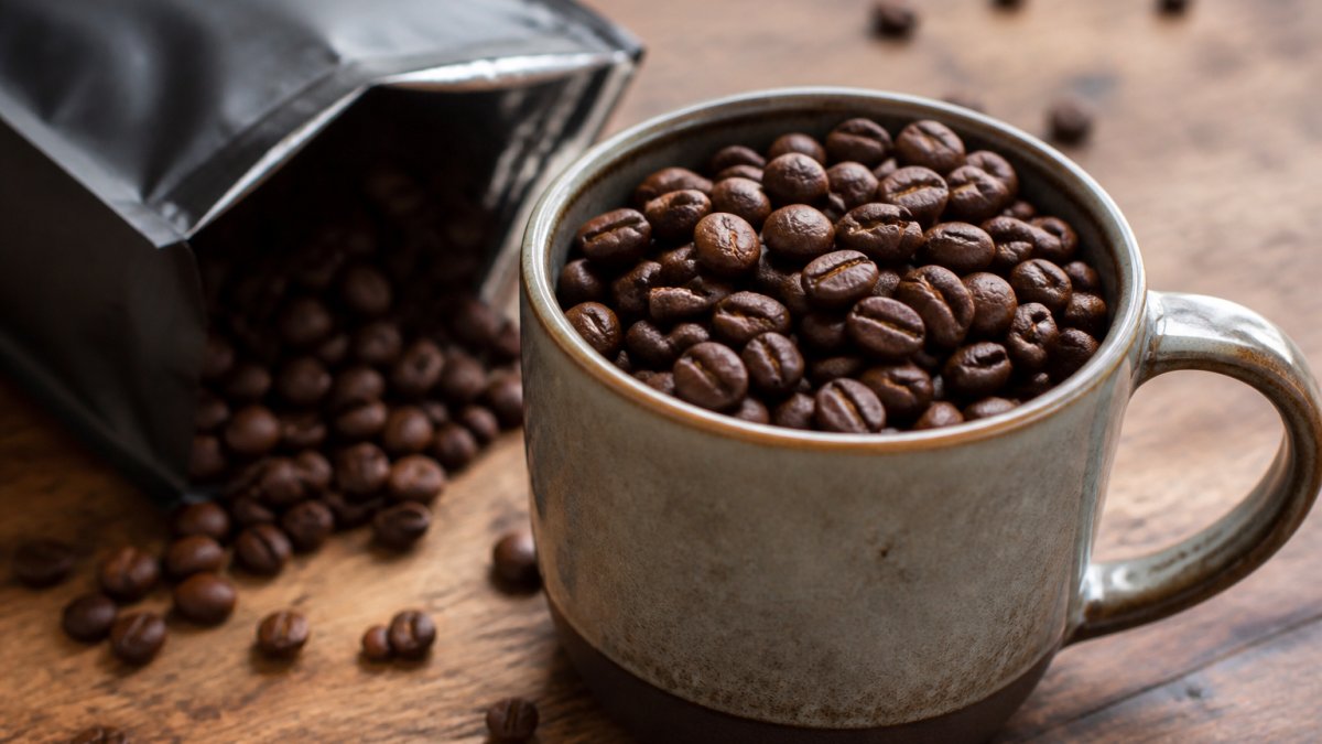 Close-up of fresh, dark roasted coffee beans in a ceramic mug with rich brown color and texture, whole coffee bag visible in background