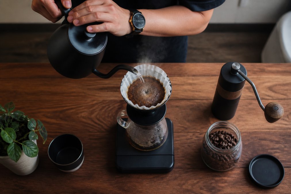 Hands pouring hot water from a gooseneck kettle over grounds to brew pour over coffee on a wooden counter