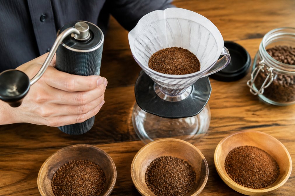 Close-up of a hand holding a burr grinder next to a pour over cone with freshly ground coffee, showing different grind textures on a wooden table