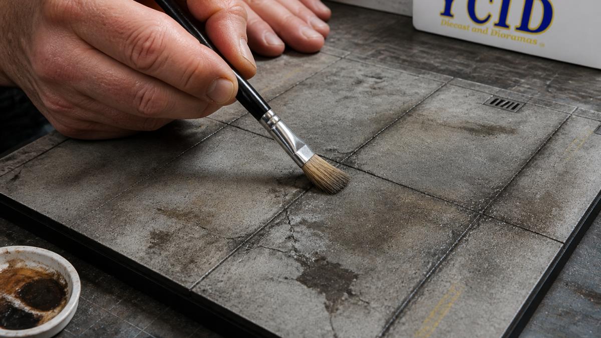 Close-up of modeler's hands applying dry brush highlights to a detailed diorama concrete surface showing realistic weathering effects, oil stains, and aged texture under bright work lighting
