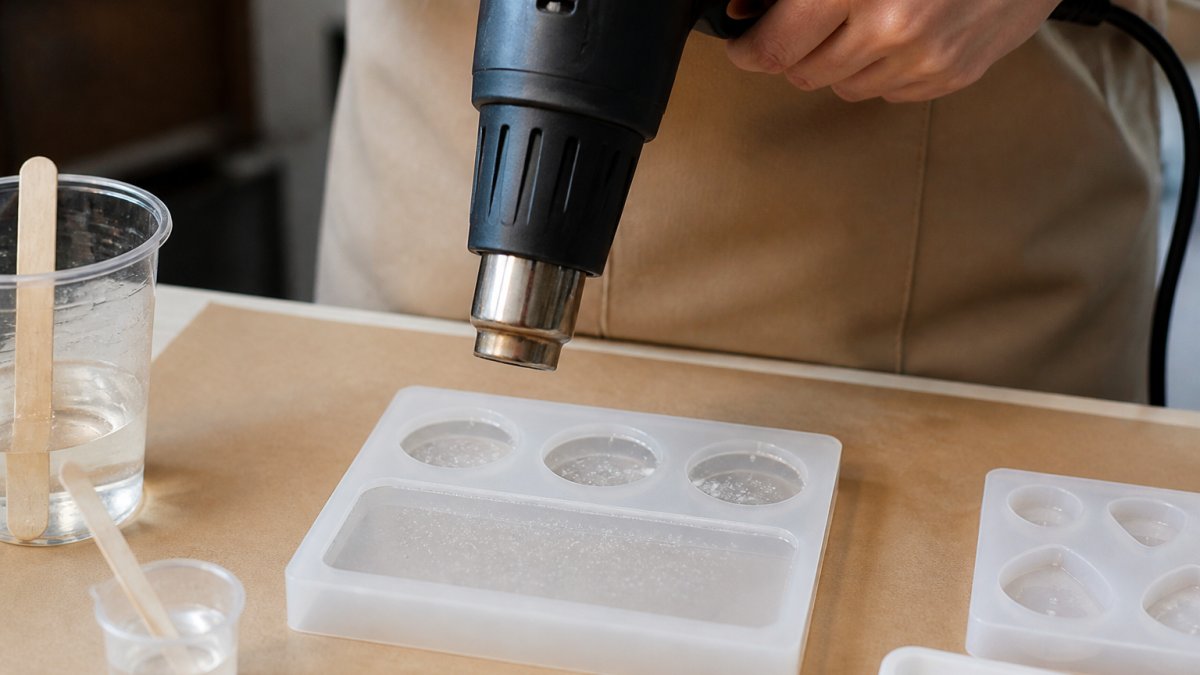 Close-up of hands holding a heat gun several inches above a silicone mold filled with clear epoxy resin, small bubbles rising to the surface, on a clean craft table with mixing cups and stir sticks nearby