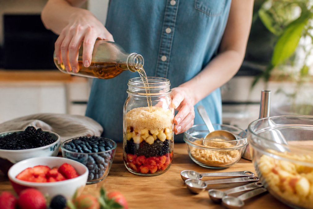 Close-up of fresh berries and diced fruit being layered in a clear glass jar with granulated cane sugar and apple cider vinegar being poured in, hands measuring ingredients with metal spoons, natural kitchen lighting
