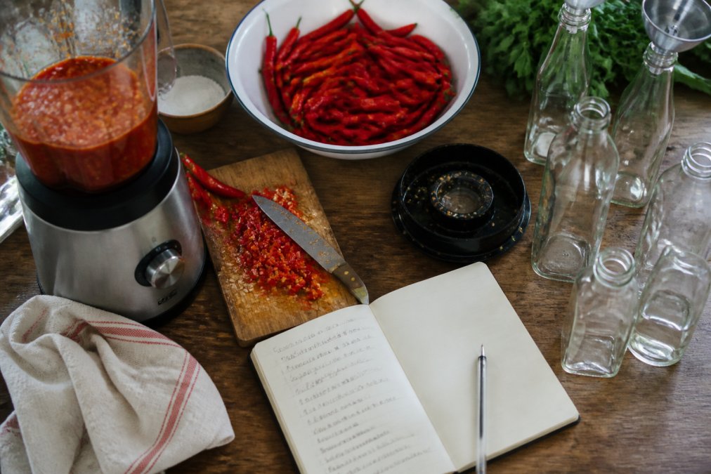 Overhead shot of a home kitchen counter with fresh red chillies, a blender, glass bottles and a handwritten notebook showing a small batch hot sauce recipe in progress