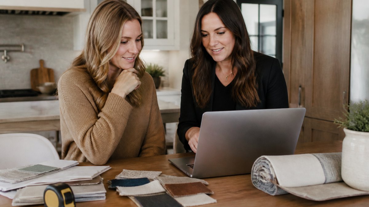 A homeowner and interior designer reviewing a renovation budget spreadsheet on a laptop at a kitchen table, with fabric swatches and a tape measure nearby