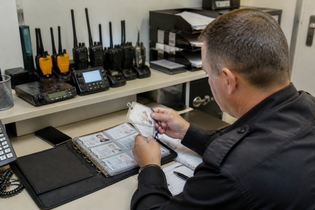 Security professional reviewing employee access credentials and ID badges at a desk with multiple monitors displaying system logs and access records