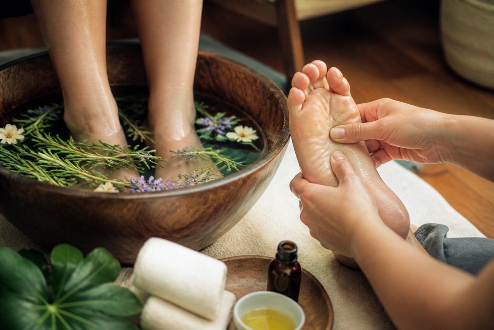 Close-up of someone's feet soaking in a warm therapeutic foot bath with essential oils and herbs visible in the water, hands gently massaging the sole in a peaceful home spa setting