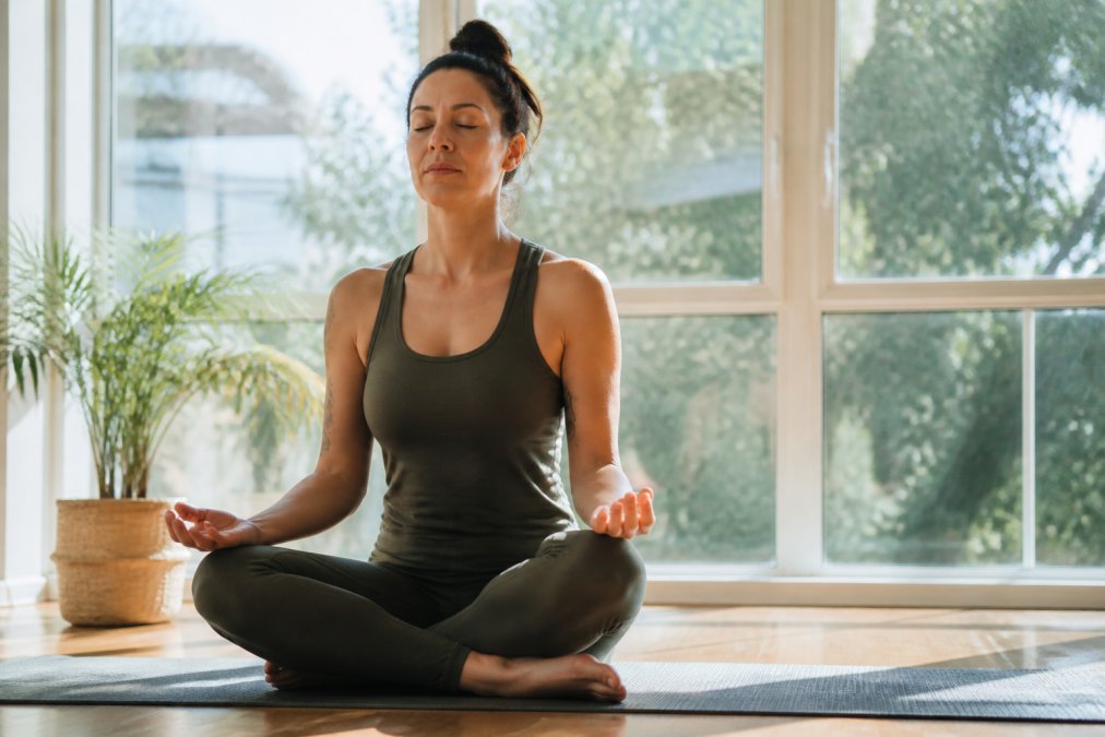 A person sitting cross-legged on a yoga mat near a sunlit window, eyes closed, hands resting gently on their knees in a calm meditation pose, soft morning light filling the room