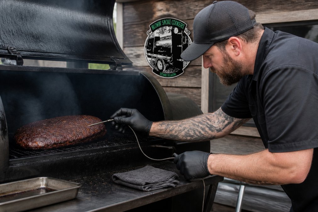 Professional pit master checking internal meat temperature with a probe thermometer inserted into a brisket inside an open smoker chamber with visible smoke, natural outdoor lighting