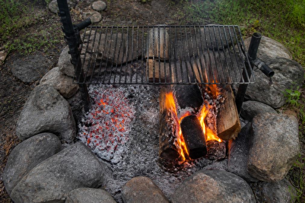 Overhead view of an active campfire with glowing coals on one side and active flames on the other, showing the distinct hot and cool zones with a cooking grate positioned above