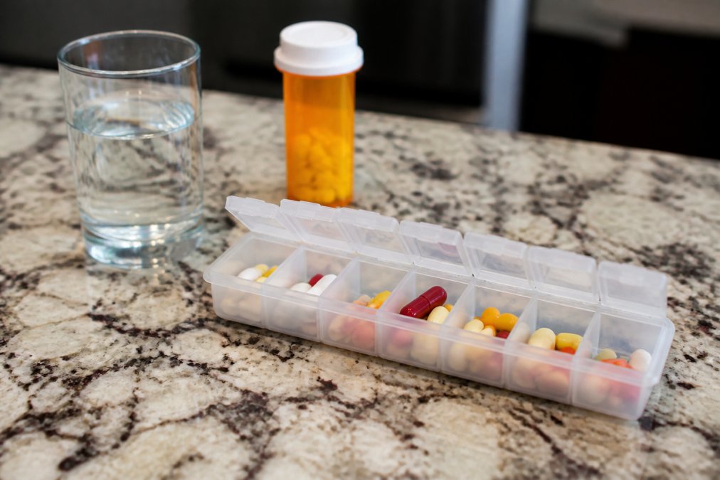 A close-up of a weekly pill organizer with compartments filled with different colored pills, a glass of water, and a prescription bottle nearby on a kitchen counter