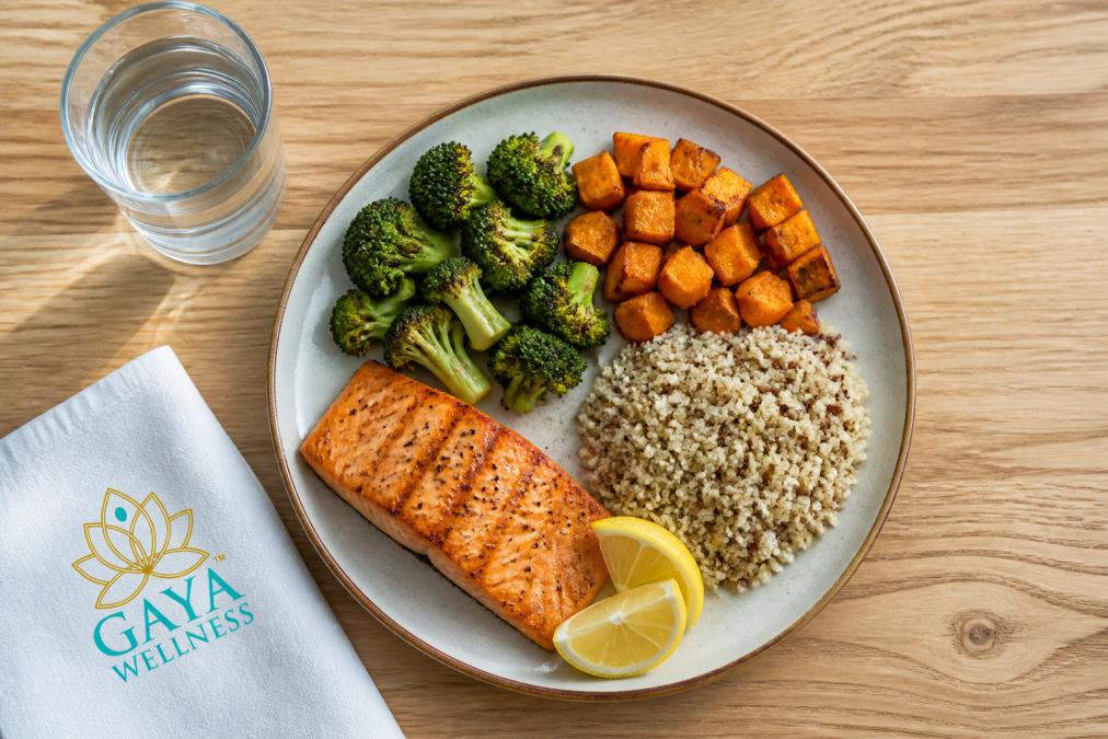 Overhead view of a colorful balanced meal plate featuring grilled salmon fillet, roasted broccoli and sweet potato, quinoa, fresh lemon wedges, and a glass of water on a natural wood table with soft natural lighting