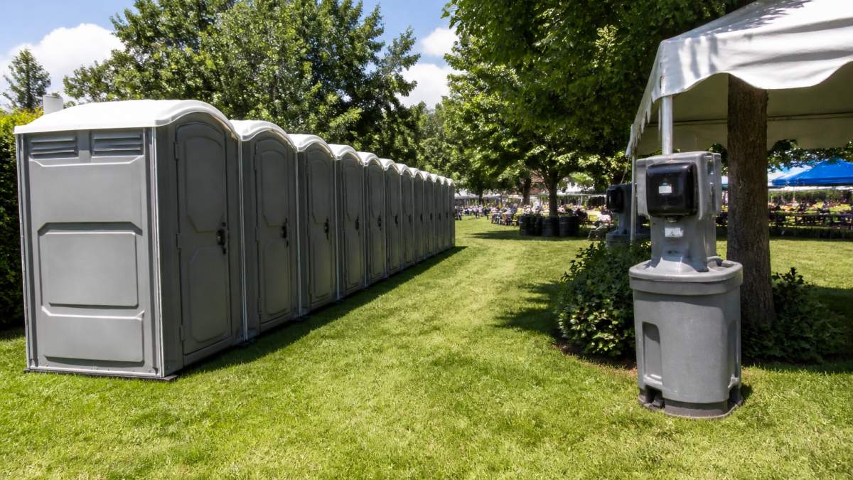 A row of clean portable toilets set up on level grass at an outdoor event, with a clear walking path and handwashing station visible nearby under daylight