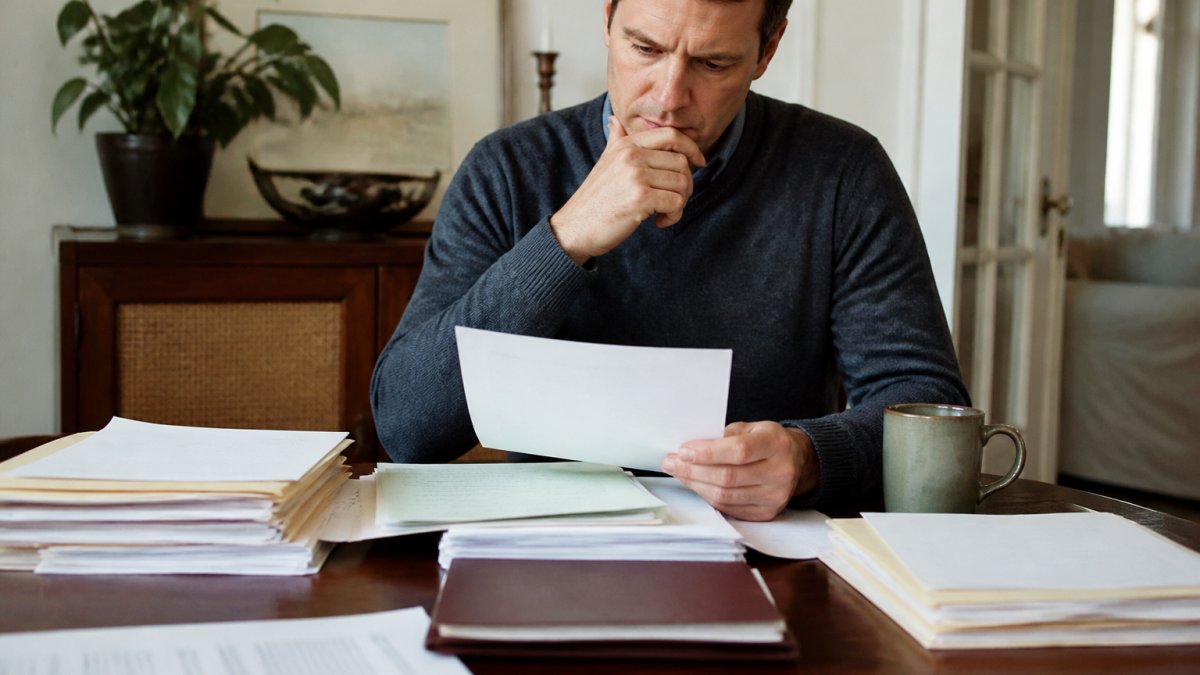 Adult child sitting at a desk surrounded by important documents, will, and estate papers, looking thoughtfully at paperwork with a cup of coffee nearby