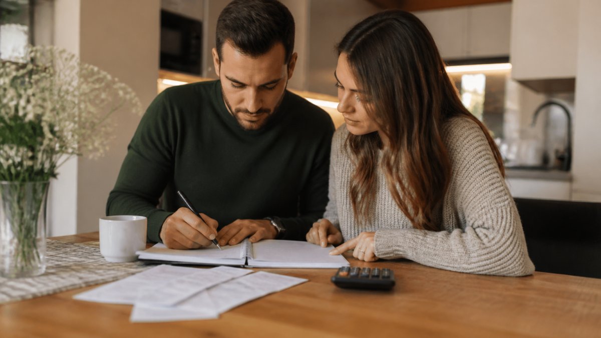 A couple sitting at a kitchen table with a laptop, a notebook, and a calculator, reviewing a wedding budget together in a warm home setting with soft evening light